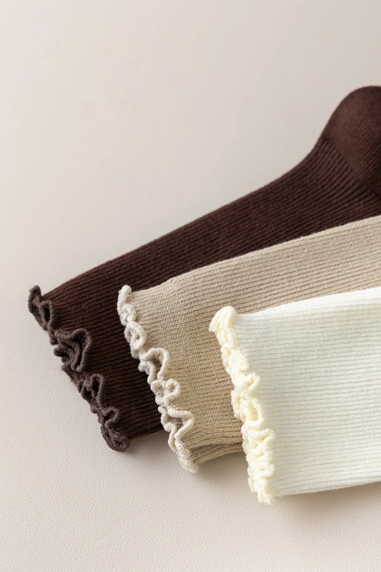Close-up of the scalloped ruffle cuff on three ruffle-edge slouch crew socks in coffee brown, ecru and bleached white.